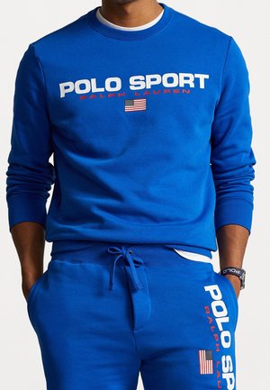 Mężczyzna noszący jasnoniebieską bluzę Polo Sport Ralph Lauren i pasujące spodnie dresowe z logo amerykańskiej flagi.