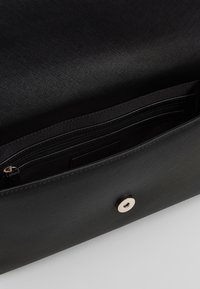 Musta teksturoitu käsilaukku, jossa sileä pinta, vetoketjullinen tasku sisällä ja metallinen nappisuljin.