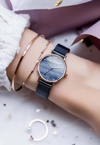Montre en or rose avec un bracelet en maille bleu foncé, dotée d'un cadran sombre et de marqueurs horaires ressemblant à des diamants. Accompagnée d'un fin bracelet en or rose.