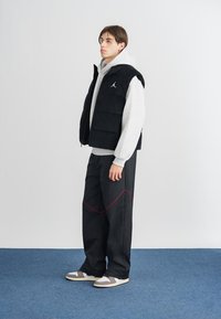 Svart pufferväst över en grå huvtröja, i kombination med svarta byxor med röd söm och grå sneakers. Släta texturer genom hela outfiten.