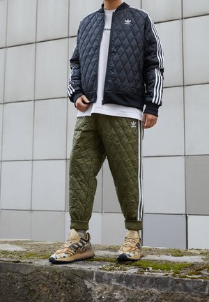 Person iført sort quiltet jakke, hvid skjorte, quiltede olivengrønne bukser og beige mønstrede sneakers, der står på en mosdækket stenoverflade.