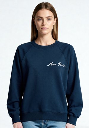 Giovane donna con capelli lisci che indossa una felpa blu navy con la scritta "Mira Paris" ricamata sul petto, in piedi davanti a uno sfondo semplice.
