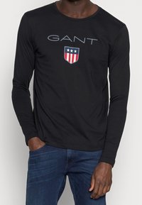 Schwarzes Langarm-T-Shirt aus Baumwolle mit rundem Ausschnitt, das den Text "GANT" und ein Wappenlogo mit roten Streifen und Sternen zeigt.