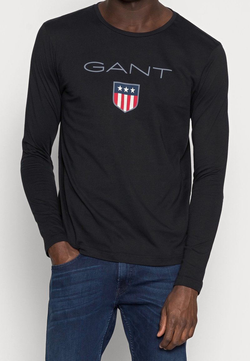 Schwarzes Langarm-T-Shirt aus Baumwolle mit rundem Ausschnitt, das den Text "GANT" und ein Wappenlogo mit roten Streifen und Sternen zeigt.