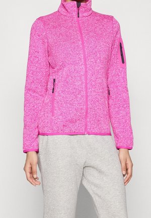 Femme portant une veste polaire rose ajustée à fermeture éclair avec des poches à l'avant et sur la manche, associée à un pantalon de jogging gris clair.
