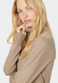 Šviesiai rudas megztinis su aukštu kaklu, pagamintas iš minkšto trikotažo, su raišteliais ant rankovių ir laisvu siluetu. Modelis pozuoja ranką laikydama prie smakro.