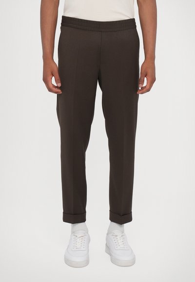 Pantalons bruns avec une taille élastique, jambes fuselées et ourlets roulés, portés avec des baskets blanches et des chaussettes grises. Texture lisse.