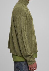 Pull en tricot vert à col roulé avec motif torsadé, coupe ample, poignets et ourlet côtelés, superposé à un t-shirt vert.
