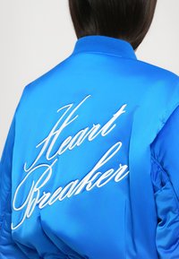 Blå satin bomberjakke med hvid broderet tekst "Heartbreaker" fremtrædende på ryggen, med ribbet krave og teksturerede ærmer.