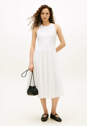 Kvinde med krøllet hår iført en hvid midikjole uden ærmer, der holder en sort snørelukket pung og har sorte flettede flade sko på.