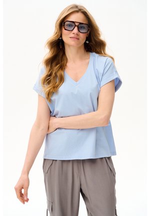 Kobieta z długimi falującymi włosami, nosząca duże okulary przeciwsłoneczne w oprawkach w kolorze żółwia, jasnoniebieską koszulkę z dekoltem w serek, szare spodnie i zwisające kolczyki.