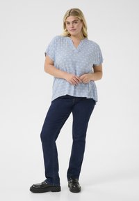 Blusa azzurra chiara a maniche corte con scollatura a V, caratterizzata da un motivo geometrico bianco. Abbinata a jeans dritti blu scuro e scarpe nere.