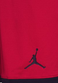 Tejido texturizado rojo con un logotipo en silueta negra de una persona saltando y sosteniendo una pelota, cosido en el borde inferior.