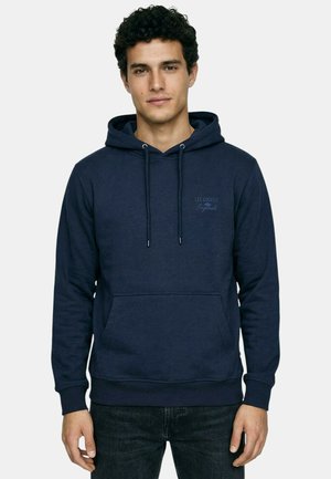 Jeune homme aux cheveux bouclés portant un sweat à capuche bleu marine Lee Cooper et un jean noir, debout devant un fond clair uni.