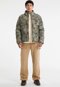 Kurtka puchowa w kamuflażu w odcieniach zieleni i brązu, z wysokim kołnierzem i zamkiem błyskawicznym, zestawiona z beżowymi spodniami typu cargo i ciemnymi butami.