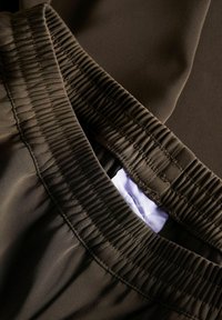 Brūna auduma ar gludu tekstūru, ar savilktu elastīgu jostu un gaiši violetu iekšējo birku. Tuvplāns uz apģērba detaļām.