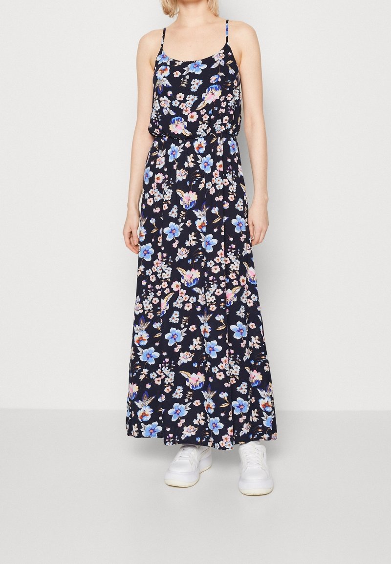 Femme portant une robe maxi noire avec un imprimé floral bleu et rose et des fines bretelles, associée à des baskets blanches, debout contre un mur uni.