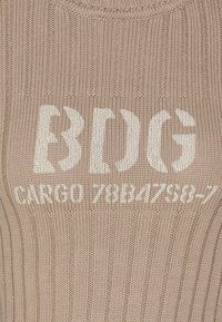Ribbestrikket topp i lys tan med hvit tekst "BDG CARGO 78B47858-7," med rund hals og teksturert stoff.
