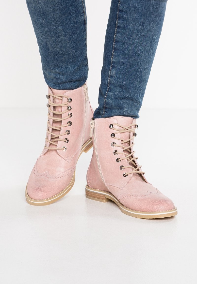 Personne portant des bottines à lacets en daim rose pâle avec des fermetures éclair latérales et un jean en denim bleu, debout sur un sol blanc.