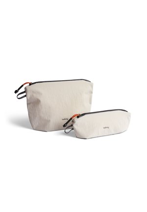 Trousse de toilette en tissu beige clair, une grande et une petite, avec des fermetures éclair noires et des accents orange. Présente une texture subtile et un logo.