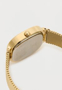 Aranyszínű, téglalap alakú óra, hálós pánttal és lekerekített koronával. Sima fémháza van, átlátszó üveglappal és ezüst színű hátlappal.