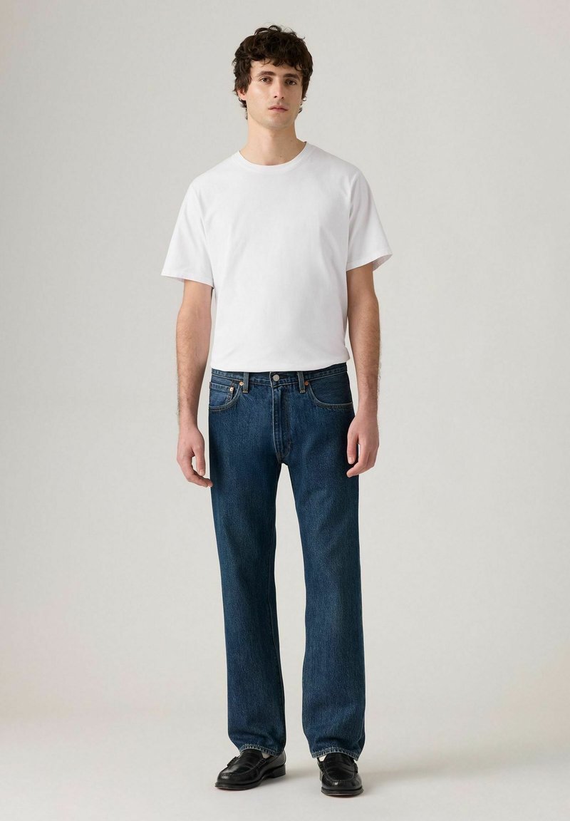 Camiseta blanca de algodón combinada con vaqueros de mezclilla azul oscuro. Los vaqueros presentan un corte clásico y un diseño de cinco bolsillos. Zapatos oscuros complementan el conjunto.