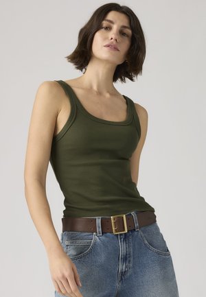 Jeune femme aux cheveux courts et foncés portant un débardeur vert olive rentré dans un jean taille haute bleu avec une ceinture marron.