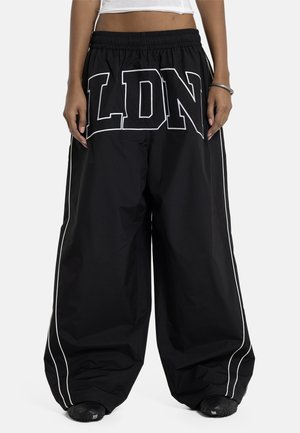 Schwarze Hosen mit weitem Bein, weißen Paspelierungen und großen "LDN"-Buchstaben über der Taille, getragen mit schwarzen Schuhen und einem weißen, kurz geschnittenen Oberteil.