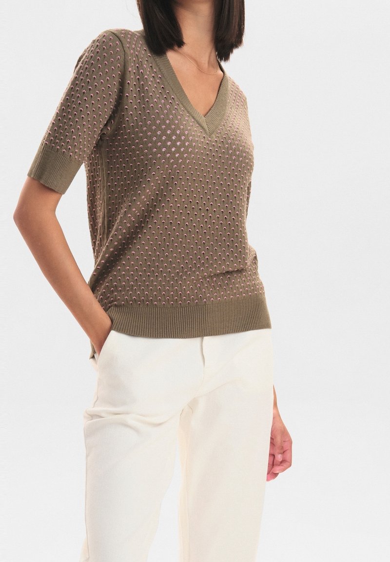 Olivgrön stickad tröja med V-ringning, korta ärmar och texturerat mönster av små rosa prickar. Ribbad nederkant och manschetter. Matchad med vita byxor.