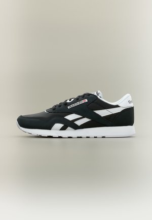 Czarno-białe buty sportowe Reebok z białą podeszwą, logo na boku, pięcie i języku, na jednolitym jasnoszarym tle.