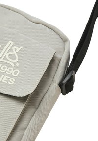 Bolso de hombro de lona gris con cierre de solapa, que cuenta con una correa ajustable negra, tela texturizada y logotipo impreso en blanco en la parte frontal.