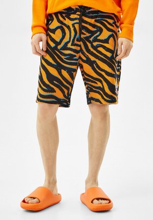 Person trägt orange-schwarze Shorts mit Tigerstreifen, einen orangen Pullover und leuchtend orangefarbene Sandalen, steht vor einem weißen Hintergrund.
