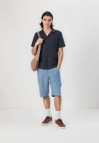 Chemise à rayures navy à manches courtes, short en denim clair, baskets brunes et sac à bandoulière beige. Design simple avec boutons et textures contrastées.