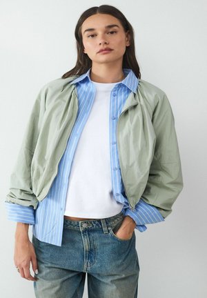 Lichtgroene zip-up jack over een blauw gestreept shirt, gecombineerd met een wit t-shirt en blauwe jeans; zachte texturen en een relaxte pasvorm.