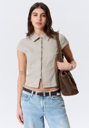 Jeune femme aux cheveux bruns mi-longs portant un haut beige à manches courtes avec fermeture éclair, un jean bleu clair, une ceinture noire et un sac à épaule marron.