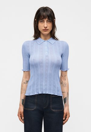 Woman with shoulder-length dark hair wearing a light blue short-sleeve knit top with a collar and dark denim jeans, standing against a white background.