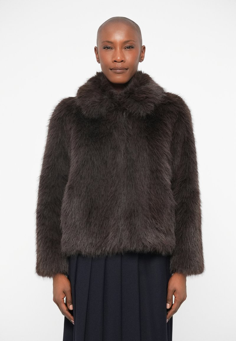 Casaco de pelúcia castanho com um colarinho alto, apresentando uma textura suave e um design curto, usado sobre uma saia plissada escura.