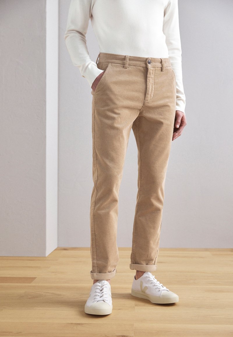 Pantalones de pana beige con pierna recta, que cuentan con cierre de cremallera y botón en el frente, combinados con zapatillas blancas.