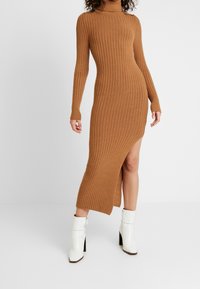 Robe en maille côtelée marron, dotée de manches longues et d'une fente sur le côté. Portée avec des bottines blanches, mettant en valeur la silhouette épurée de la robe.