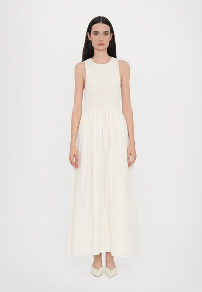 Robe longue blanche avec un corsage smocké ajusté et une jupe fluide. Le tissu est lisse, avec un col rond et un design sans manches.