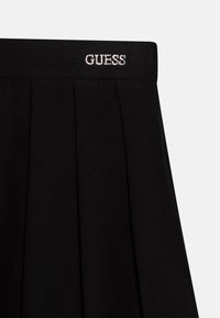 Jupe plissée noire avec une texture lisse et une ceinture portant le logo "GUESS" imprimé en strass. Design minimaliste avec une silhouette classique.