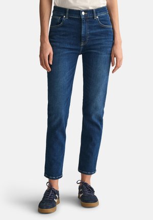 Donkerblauwe denim jeans met een aansluitend ontwerp, vijf zakken en een rechte pijp. Draagt een paar marineblauwe sneakers met tan accenten.