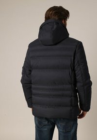 Homme portant une veste à capuche rembourrée noire et un jean bleu, vu de dos sur un fond uni.