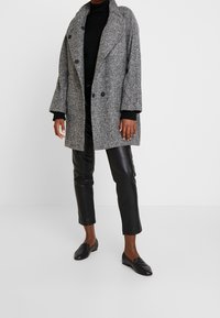 Grijze houndstooth jas met brede revers, dubbele voorknopen en zwarte gebreide accenten. Gecombineerd met zwarte leren broek en platte schoenen.