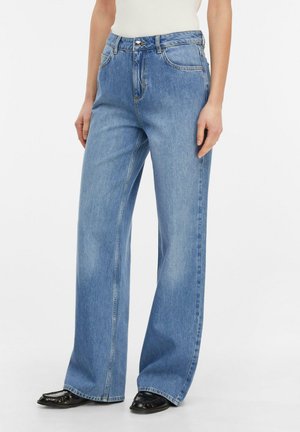 Osoba ubrana w jasnoniebieskie, szerokie dżinsy z pięcioma kieszeniami oraz czarne buty na kwadratowym nosku, stojąca na jednolitym tle.