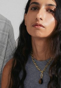 Gold chain necklace featuring a teardrop pendant with amber tones, worn over a gray sleeveless top. Long dark hair frames the face.