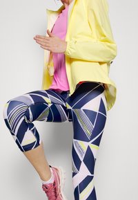 Athletic wear featuring a yellow lightweight jacket, pink top, and navy patterned leggings with geometric shapes in white and neon accents.