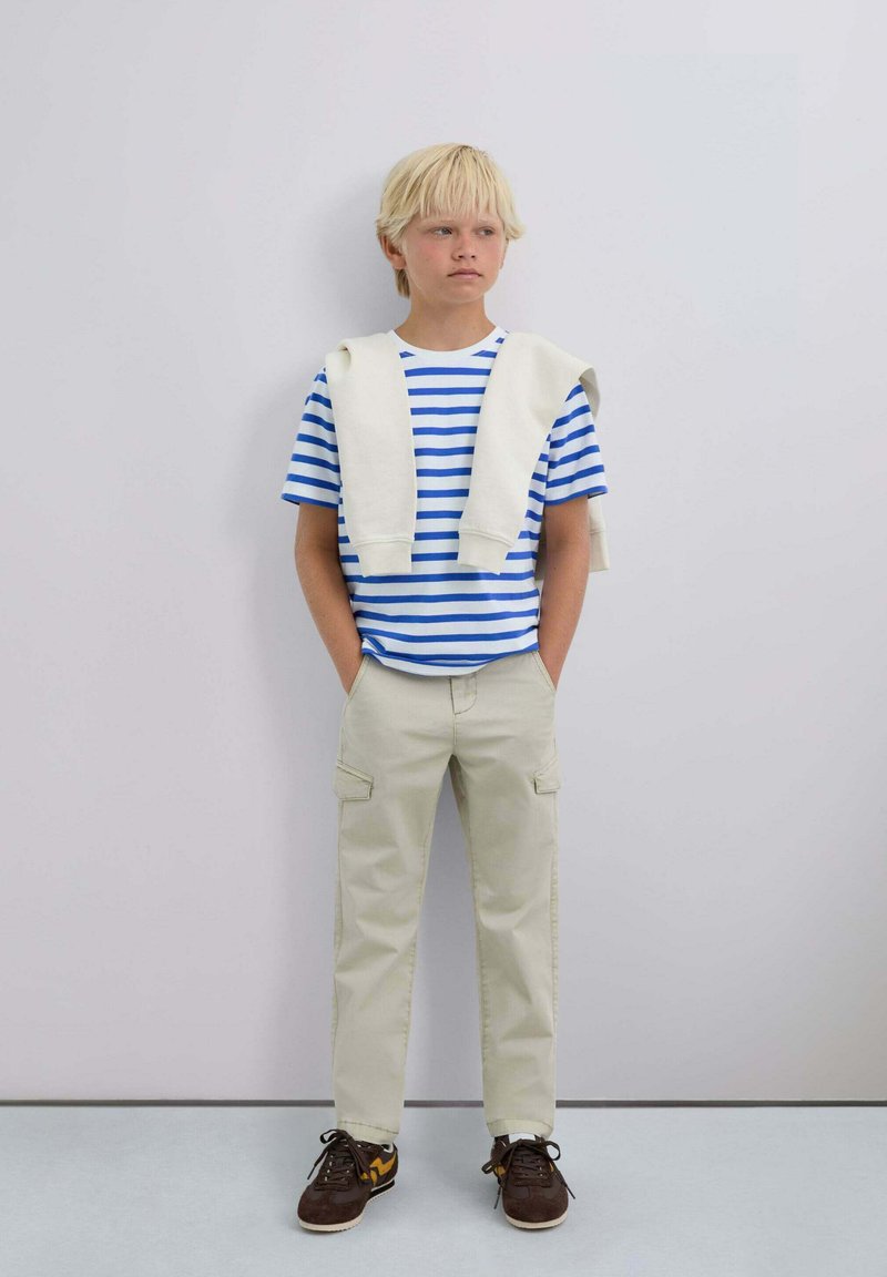 Camiseta a rayas azul y blanca, suéter crema sobre los hombros, pantalones cargo beiges y zapatillas marrones con acentos amarillos.