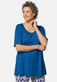 Türkises Kurzarmoberteil mit Rundhalsausschnitt und Raffung, getragen über floral gemusterten Hosen. Glattes Material mit lockerer Passform.