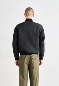 Pull noir texturé avec un dos côtelé, associé à un pantalon vert olive avec des poches arrière et une étiquette bleue. Vu de dos.